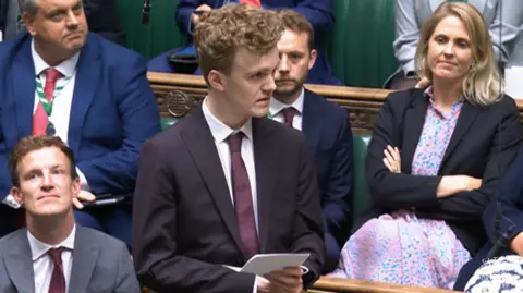 House of Commons Sam Carling, wearing a dark suit, white shirt and maroon tie, stands in the House of Commons. He is holding a white piece of paper. Behind him, sitting on benches, are other MPs.