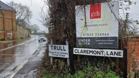 Dawn Johnson A sign reading 'Trull please drive carefully through village' with a much larger estate agents' board next to it advertising building plots