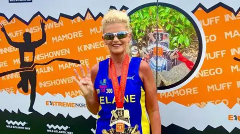 Elaine McAnulty A woman with blonde haired tied up, wearing sunglasses in front of an orange marathon banner. She is wearing a blue and yellow vest and has a large medal around her neck, on a red lanyard. She is holding four fingers up and smiling.
