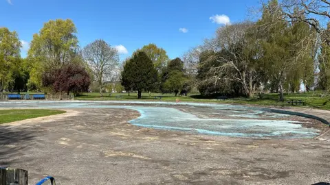 The drained paddling pool at Pickering Park it is grey concrete with a patch of blue in the middle and is surrounded by tall trees
