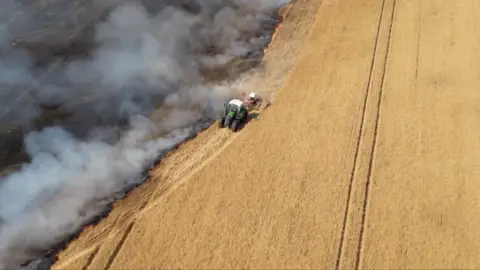 A drone shot of a tractor driving in a field separating a fire from untouched crops.
