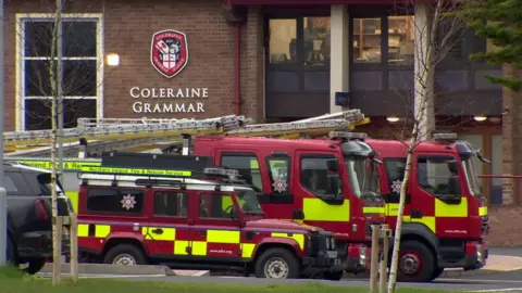 Three red and yellow hi-viz fire trucks sit infront of a red brick building which has the words "Coleraine Grammar" on it, in silver writing.
