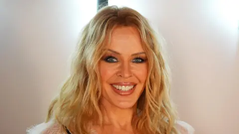 Head-shot of a woman with wavy shoulder-length blonde hair. She is smiling at the camera against a light background.