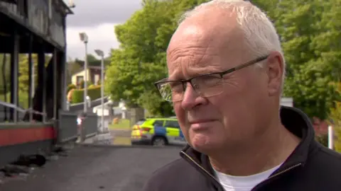 A man with white hair and glasses is standing outside outside a car showroom that has been damaged with fire and smoke. A police car is in the background of the image. 