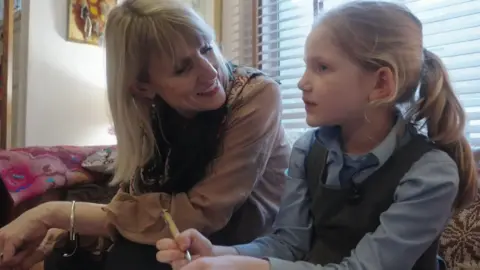 Dr Dunlop sits next to a primary-aged schoolchild in uniform while the girl studies Romanian. The child is holding a pen and is thinking 
