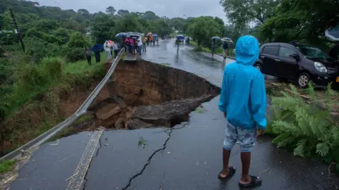 AFP/Getty Images A child wearing a blue raincoat looks on at a collapsed road. A fallen tree lies beside the child and people holding umbrellas huddle in the distance.