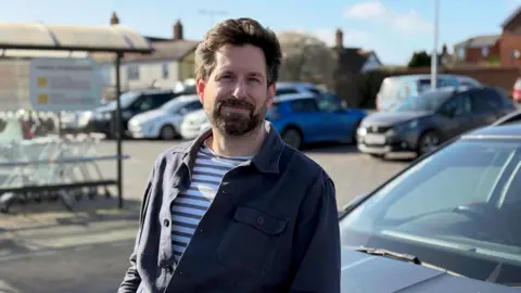 Gareth Wild stands in front of his car at the car park of his local supermarket. He looks at the camera and has dark brown hair and a beard. He is wearing a striped t-shirt underneath a navy blue shirt.