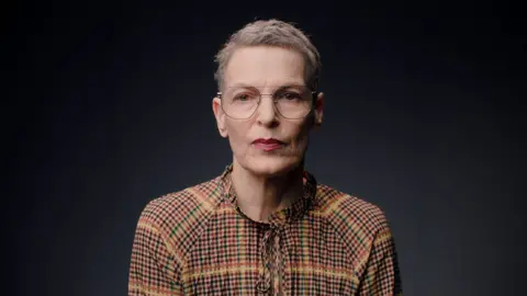A woman with short grey hair, large thin-rimmed glasses and wearing a checked top looks ahead.