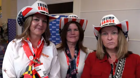 Three middle-aged women standing side by side wearing Trump Vance cowboy hats