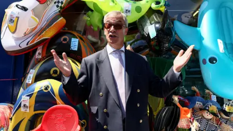 Reuters Nigel Farage wears a suit and sunglasses. He's standing with his arms stretched to his side, in front of several inflatable toys
