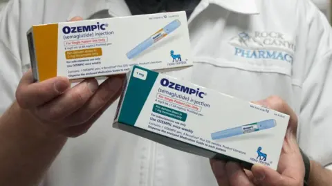 A pharmacist holding two boxes in each hand of Ozempic, showing different doses (one 2mg and another 1mg). The boxes say Ozempic (semaglutide injection) and feature a photo of the inkection pen, as well as instructions on how to use the drug. They also feature the logo of manufacturer Novo Nordisk.