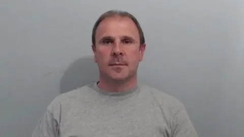 A mug shot of Irvine. It is a head and shoulders image, he is wearing a grey sweatshirt and has short brown hair. 