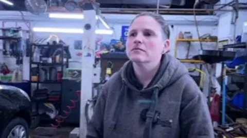 Rachael Longstaff pictured inside the garage workshop. She is wearing a grey hoodie and has tied back hair. There are shelves with tools in the background and a car on a ramp.