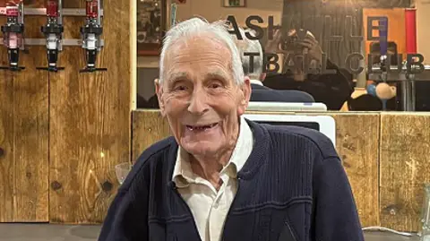 Ashville FC John Dennett with white hair and wearing a navy cardigan over a cream shirt. He is standing behind the bar at Ashville FC with a mirror behind him with the club's name engraved on it. He is smiling.