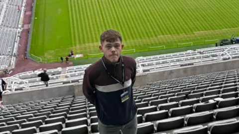 Pardoe Kendrick wearing a dark top, he is high up in the stands of a football stadium with the pitch in the background.