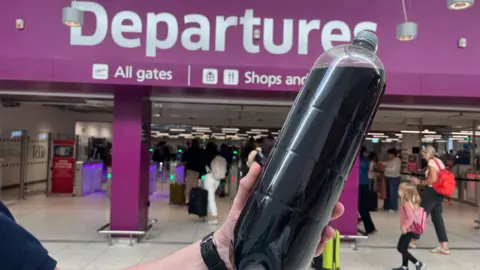 A 2 litre bottle in front of a purple sign that reads "Departures". People walk through the opening below the sign.