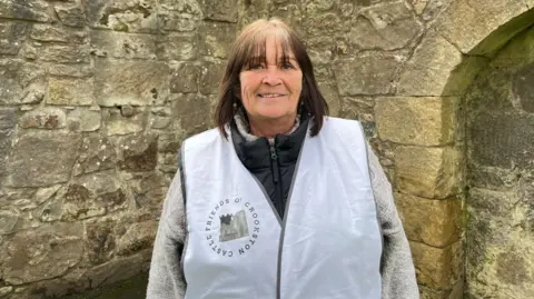 Mary-Ann Williamson, a woman with short dark hair and wearing a white bib saying Friends of Crookston Castle, standing in front of a castle wall