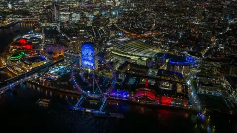 jasonhawkes.com An aerial view of the Thames featuring the a large ferris wheel and railway station lit up in pink 
