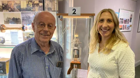 Rod Viveash with short white hair and glasses wearing a shirt of small blue and white squares and Carrie with long blond hair wearing a white top. They are standing in front of an old glass value in a display case numbered as 2. There is a larger display case behind them.