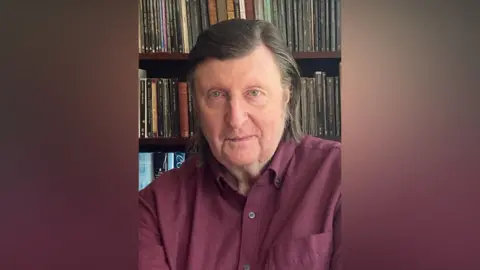 Robin Page A man with slicked back, shoulder-length brown hair, with a parting on the left. He is looking at the camera and smiles slightly. He is in front of a shelf filled with CDs. He is wearing a dark red collared shirt. 