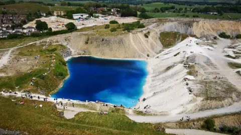 A blue pool of water surrounded by a green and brown quarry with people on the edge, picture of the Blue Lagoon taken from a BBC story in 2020