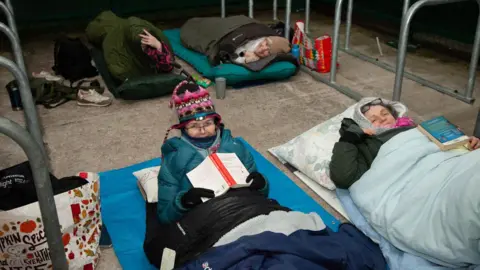 Threshold People in sleeping bags wrapped up warm and smiling at the camera. It is a cold looking night and a couple of the women hold books.