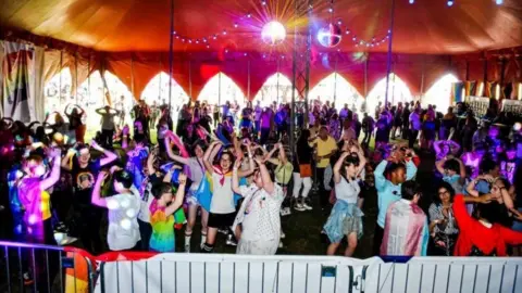 Herts Pride A large crowd gathers under a decorated tent, dancing beneath colourful lights and disco balls at a recent Herts Pride event.