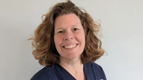 Royal Cornwall Hospitals NHS Trust Hermione Bigwood smiles while wearing dark blue medical scrubs on. She has shoulder-length wavy hair which is light brown.