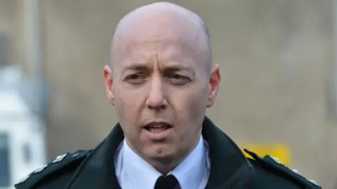 Pacemaker A bald man wears a dark green jacket with shoulder straps. Under the jacket he has a white shirt and black tie.