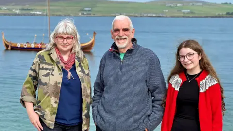 Three people in warm woollies in front of a bay with a viking style longboat in it.