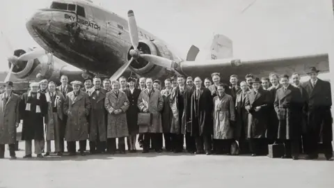 A black and white picture of about 30 men in front of an aircraft which has a propeller and one of its wings in shot. One air hostess can be seen in the front of the photo, linking arms with two of the passengers. The men are mainly wearing long coats and some are wearing flat caps or bowler hats. 