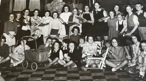 Ann Mathers A group of children gathered in the town hall with various toys.
