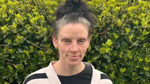 Afton Southern A woman called Afton Southern stands in front of a bush. Her greying black hair is tied into a top knot. She has a black and white stripey cardigan on and a black T-shirt underneath.
