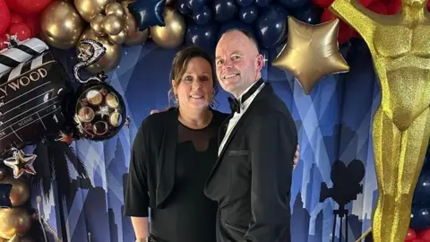 Family A woman with long brown hair and a black dress next to a man with grey hair and a grey beard in a dark suit in front of a colourful background with a large gold statue beside them