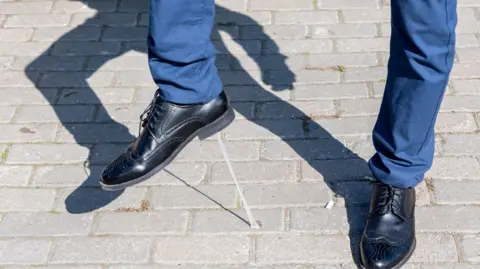 A stock photo showing the bottom half of a pair of legs in blue trousers and smart black shoes. The person is lifting up their right foot - they have stepped in gum, which is now stuck to the bottom of their shoe.