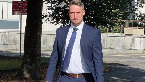 Huw Orphan, wearing a blue suit, white shirt and tie and carrying a bag, looks at the camera as he walks near Cardiff Crown Court