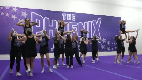 A group of cheerleaders are practising in a hall with purple flooring and a purple banner on the wall. The trainees are all wearing black and are holding some of the people up in the air.