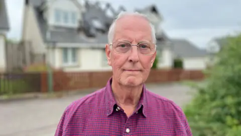 Stuart Drummond standing in front of his damaged house wearing a pink shirt and glasses. 