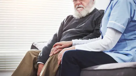 Getty Images A stock image of a female nurse in a blue uniform holding the hand of an elderly man while they are sat on a sofa.