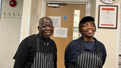 Two older smiling charity volunteers, Webster Modest on the left has short cut grey hair and wears a dark blue polo shirt under a black v-neck jumper, and a black white striped apron. Pauline Simmonds on the right is smiling broadly has short brown hair visible under and is wearing a blue baseball cap. Pauline too wears a black and white striped apron, over a blue and white pattern top, and a navy blue shirt style top.