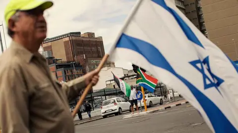 A man is seen holding up an Israeli flag as another person is seen holding South African and Palestinian flags in the background