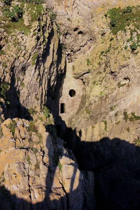 Nicole Barley Stone structure with small round and rectangular openings built into a steep, rugged cliff face. Sunlight highlights the rough beige rock, while deep shadows fall into the narrow gorge below and sparse greenery clings to the rock surface.