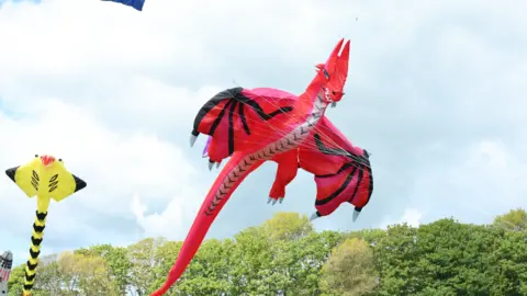 A large red kite is mid-air and designed to resemble a dragon. In the background are tree tops and a second yellow kite.