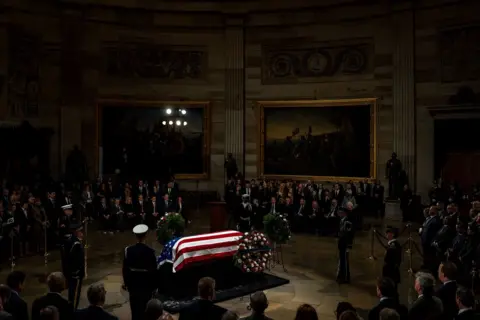 Reuters A coffin draped in an American flag surrounded by wreaths, guarded by military personnel and a crowd of mourners. There are paintings on the wall of a grand, darkened room.