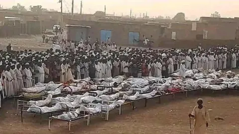 A large group of mourners stand in an open field beside a number of bodies wrapped in cloth.