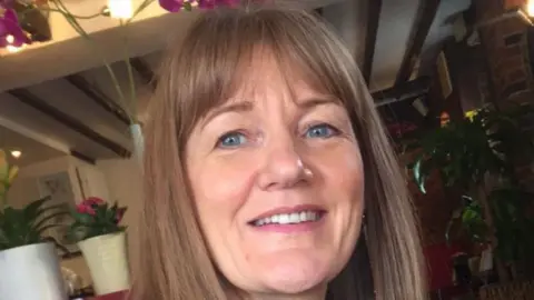 John Manitara Gillian smiles at the camera in a photograph taken before her condition progressed. She has long dark hair and a fringe and blue eyes. There are a number of plants behind her. 