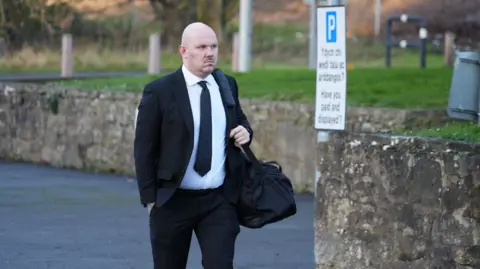 Miles Cross arriving at the court wearing a suit and a black tie. He has a black bag which he is carrying on his shoulder.