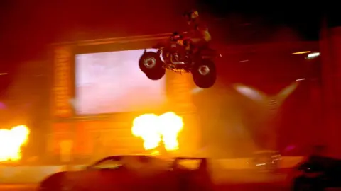 Getty Images A stunt rider on a quad bike jumps over a car in an arena, with flares of fire surrounding him. 