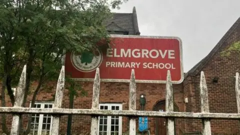 BBC Picture is taken from behind a white gate. Behind the gate is a red sign which says 'Elmgrove Primary School', to the right of the sign is a green tree. Behind the sign is a red brick building with white window panes.