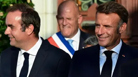 EPA/Shutterstock Sébastien Lecornu, who has dark hair and is wearing a dark suit, smiles as he looks off to the side while standing next to French President Emmanuel Macron, who is also smiling and wearing a dark suit.